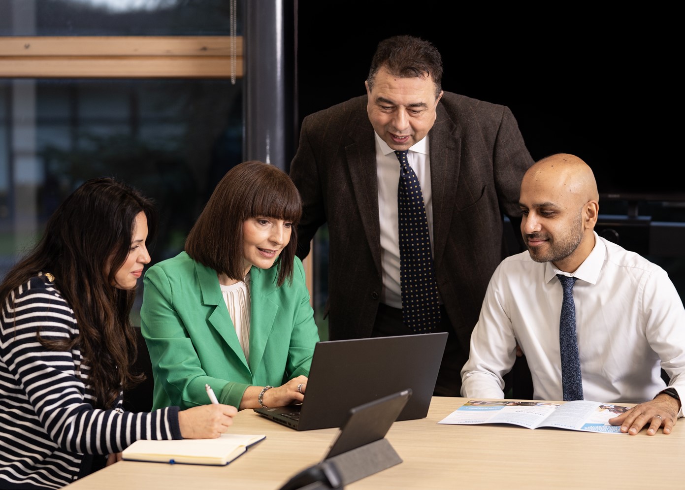 Four people are gathered around a laptop. There are two women sat at the table, and two men. One is sitting and the other is standing bending down to the laptop