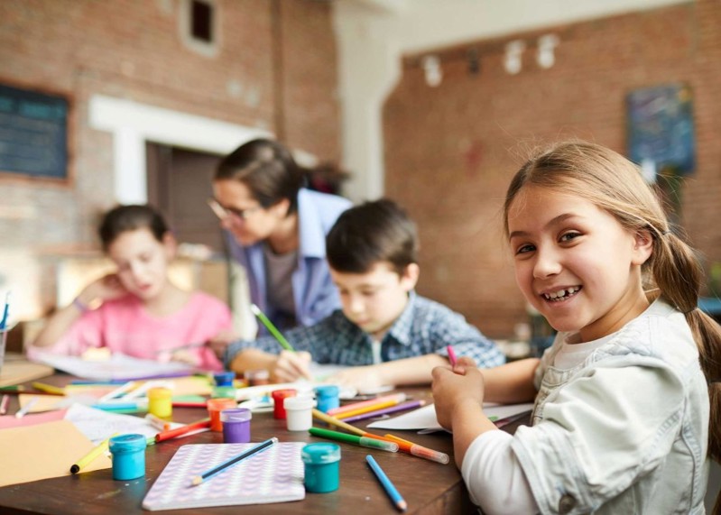 Three children sit at a desk drawing and painting. One girl is turned to the camera, smiling.