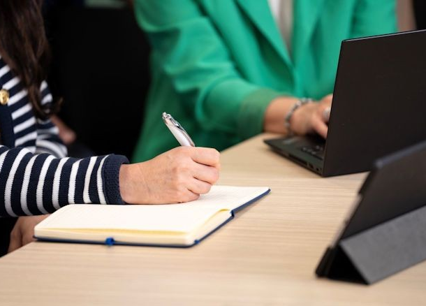 Two people sat at a desk. One is typing on a laptop, and the other writing in a notepad